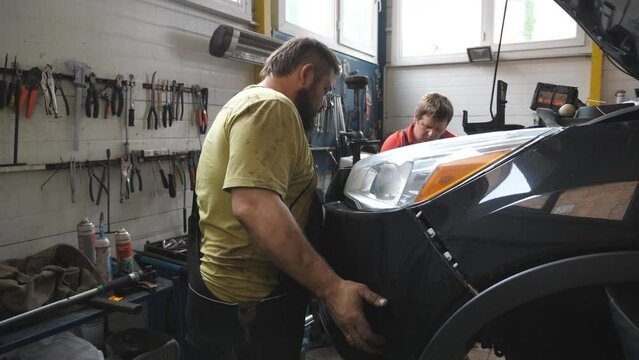 Two mechanics removing front bumper from car for repair in auto service. Professional repairmans dismantling automobile before fix at workshop or garage. Concept of vehicle maintenance and diagnostic