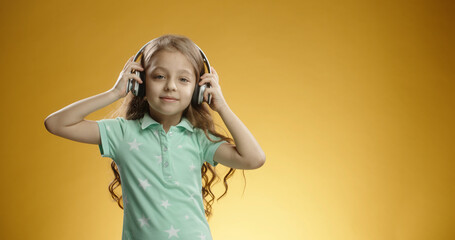 Portrait of beautiful little girl wearing blue t-shirt and headset, listening to the music, isolated on yellow colored background. Copy space