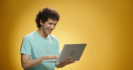 Joyful asian guy with curly hair using his laptop, typing on keyboard and happily smiling, isolated on yellow background - close up shot 