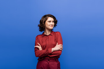 cheerful woman with short curly hair standing with folded arms isolated on blue.