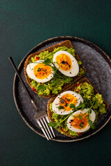 Sandwiches with rye bread and guacamole, with boiled eggs and microgreens of peas and radishes and sesame seeds, a plate with sandwiches close-up on a green background