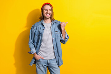 Photo of friendly guy with long hairdo dressed jeans shirt look directing empty space hand in pocket isolated on yellow color background