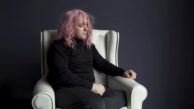 A balding man in a pink wig has a headache, he takes off his wig. A man sits in a white chair.
