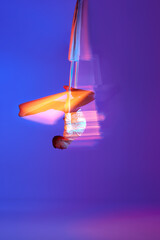 Acrobat, professional aerial gymnast hanging upside down on aerial silk against gradient blue purple background in neon with mixed lights. Concept of art, sportive lifestyle, hobby, action and motion