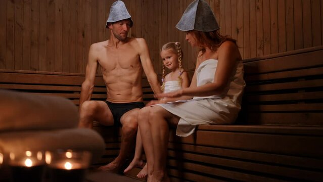 Wide shot carefree daughter with joyful father and mother in Finn sauna indoors. Positive Caucasian man woman and girl talking tickling child laughing. Relaxation and leisure concept