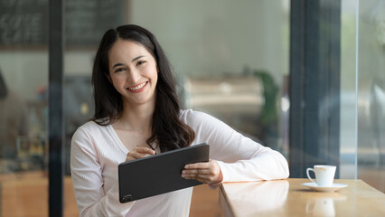 Portrait of young happy asian woman holding digital tablet and smiling at camera while standing in a cafe.