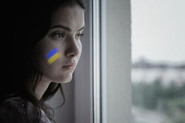 Sad brunette with ukrainian flag on face looking through window glass standing home side view....