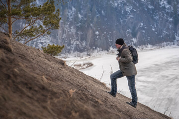 A backpacker climbs uphill on foot in winter or early spring, against the backdrop of hills and a frozen river. Outdoor recreation, solitude and hiking