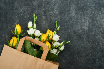 Easter concept. Bouquet of fresh white and yellow roses in paper bag on old dark concrete background. Passover Pesah celebration concept. Concept of mothers, womens day, spring background. Top view.