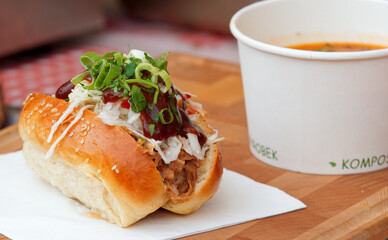 A roll or bun stuffed with pulled meat and vegetables with bowl of soup in the background at farmers market.
