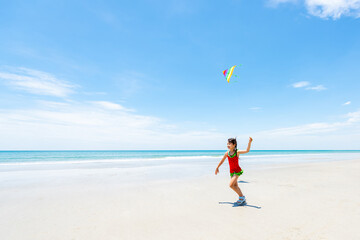 Little Asian child girl in swimsuit running and playing kite at tropical island beach in sunny day. Children kid have fun outdoor lifestyle activity travel ocean with family on summer holiday vacation