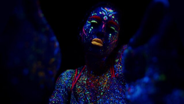 Young female performer with a fluorescent body painting stretches out her hands and performs her mystery show in a dark room highlighted by the ultraviolet close up