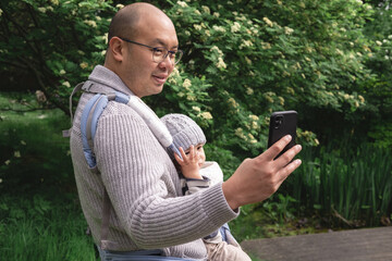 A smiley new father carrying his male infant with a baby carrier taking a selfie photo with his cell phone in nature at the park in spring season. British multicultural and multiethnic family.