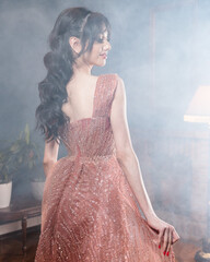 A woman in a pink dress stands in front of a vintage desk and looks into the camera.
