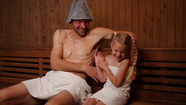 Cheerful Caucasian father tickling daughter laughing sitting in wooden Finn sauna indoors. Portrait of happy positive man and girl enjoying leisure in SPA having fun