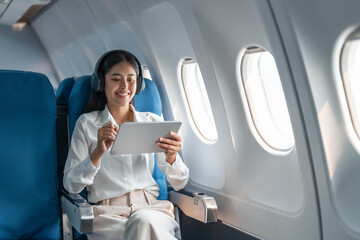 Fototapeta premium Young Asian business woman or female passenger wearing wireless headphone and working with tablet during the flight