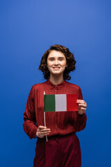 Obraz premium happy language teacher smiling while holding flag of Italy and looking at camera isolated on blue.