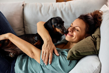 A female owner smiling while his dachshund dog licking her face, lying at home.