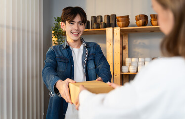 The man who owns the vase shop smiles and offers the product box to the customer. Woman receives the parcel box from her staff after packing the goods as ordered in online sell and delivery concept.