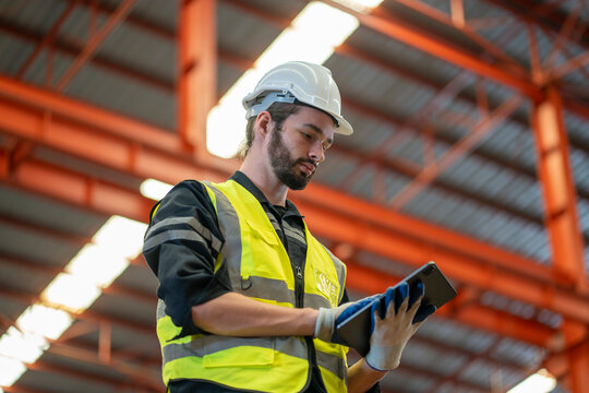 Low angle view of a worker wearing reflective jacket holding digital tablet standing in factory warehouse