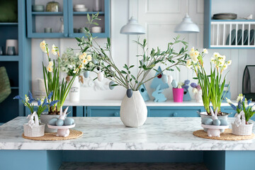 Daffodils and plants in pots, branches tree olive with easter eggs in vase. Stylish kitchen interior with easter decor. Bouquet of flowers in vase, ceramic plate with easter eggs and utensil on table 