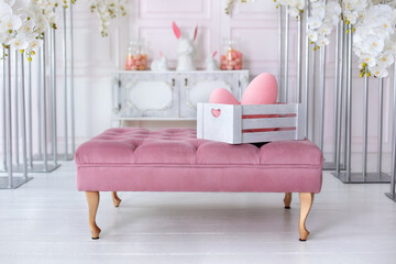 Festive Easter decoration of living room. Elegant white living room interior with pink sofa pouf and box with easter eggs. Easter festive decoration with ceramic rabbits and colorful eggs.