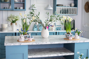 Daffodils and plants in pots, branches tree olive with easter eggs in vase. Stylish kitchen interior with easter decor. Bouquet of flowers in vase, ceramic plate with easter eggs and utensil on table	