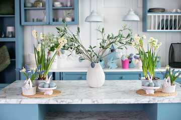 Home interior with easter decor. Kitchen utensils on table. Blue kitchen interior with furniture. Stylish cuisine with flowers in vase. Daffodils in pot and branches tree in vase with easter eggs. 