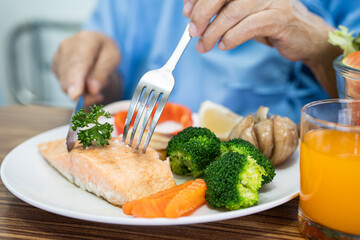 Asian senior woman patient eating Salmon steak breakfast with vegetable healthy food while sitting and hungry on bed in hospital.