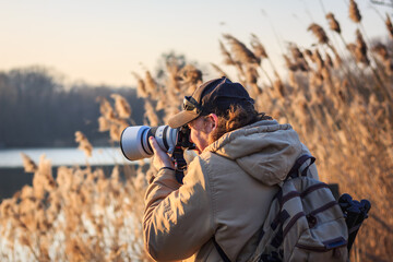 Photographer with camera hiding behind reeds at lake and photographing wildlife. Leisure activity...
