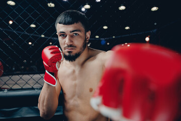 Portrait man MMA fighter with red boxing gloves trains in gym, cage octagon dark background