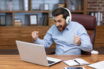 Mature businessman in shirt and headphones watching sports match at workplace inside office, man cheering having fun at work, watching video on laptop.