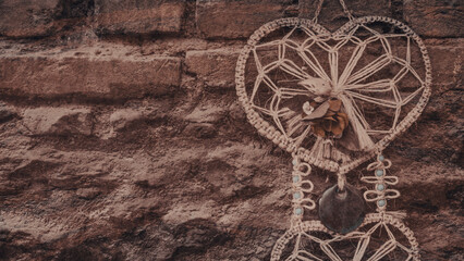 Fototapeta na wymiar A bohemian heart shaped wall hung macrame dreamcatcher. It has a rose, beads and devil eye on it. Vintage autumn colored wallpaper. copy space