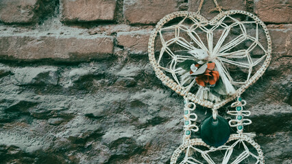 A bohemian heart shaped wall hung macrame  dreamcatcher. It has a rose, beads and devil eye on it.  Vintage autumn colored wallpaper. copy space
