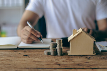Young man doing savings account with piles of coins against wooden house on wooden table saving money to buy a house home loan finance concept