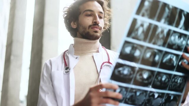 Portrait of concentrated arabian man muslim doctor therapist thinking about diagnosis while looking at results patient MRI or CT scan procedure and looking at camera at modern hospital centre outdoors