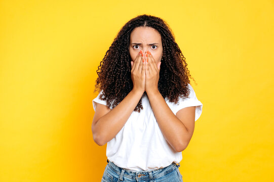 Terrified shocked brazilian or hispanic curly haired woman, covers mouth with hands, experiences fear, shock, discouragement, looks at camera, stand on isolated yellow background