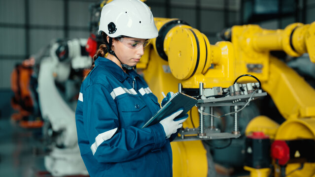Caucasian mechatronics engineer is using a tablet to record the results of examining a lot of robotic arms. A female industrial worker with responsibility for quality checking machines in the factory.