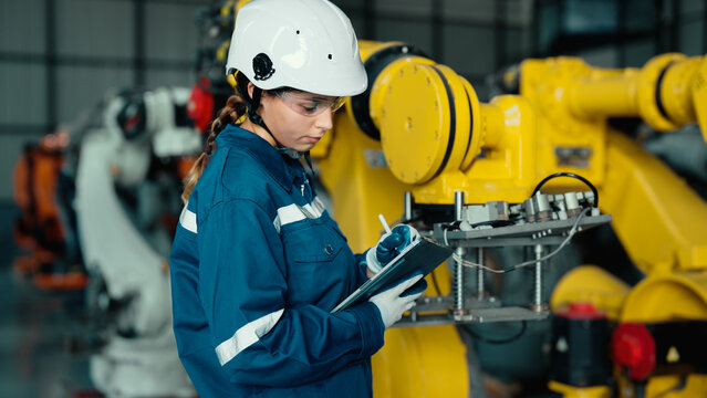 Caucasian mechatronics engineer is using a tablet to record the results of examining a lot of robotic arms. A female industrial worker with responsibility for quality checking machines in the factory.