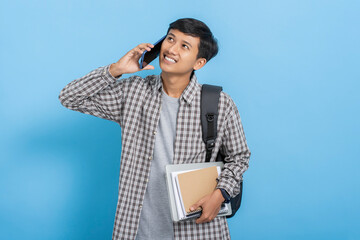 Young asian college student carrying books and backpack talking on the phone and smiling isolated over blue background