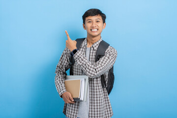 Young asian college student carrying books and backpack pointing with finger on blank copyspace isolated over blue background