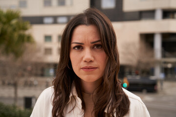 Cute serious young Caucasian girl looking at camera outside out of focus block of apartments background. Earnest female sad people with disapproving gesture. Close up of worried woman. 