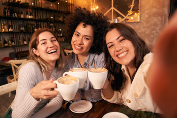 Group of cheerful female friends take a selfie toasting cups of cafe in coffee shop. Smiling and happy young girls looking at camera and having fun together. Women active on social networks.