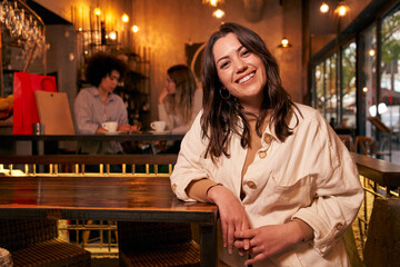 Fototapeta na wymiar Copy space portrait of cheerful girl leaning on bar table. Empowered Latin woman poses looking at smiling camera. Girlfriends having fun indoors. Three co-workers after shopping. Happy people.