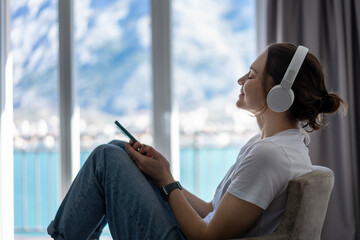 Young pretty cheerful woman listening to music in headphones sitting at home enjoying using app in smartphone