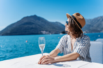 Young pretty cheerful woman in a hat enjoying a glass of champagne on the shore of a lake in the mountains during summer holidays - obrazy, fototapety, plakaty