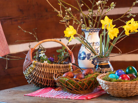 Easter and spring still life with wicker baskets, coloured decorated easter eggs ,moss, feather, vases flowers with narcissus on rustic wooden table in log cabin. Happy Easter holiday card