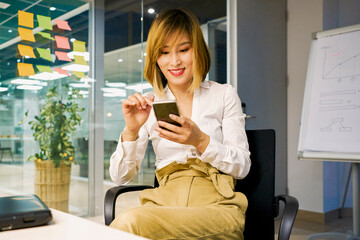 female entrepreneur in elegant outfit sitting in office chair and using smartphone in modern workspace