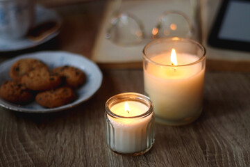 Cup of tea or coffee, plate of cookies, books, e-reader, pencil and lit candles on the table. Selective focus.