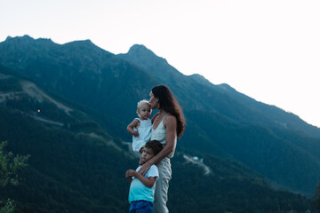 Naklejka premium a young mother with 2 children in an embrace against the backdrop of beautiful mountains and nature. The concept of rest and travel with children